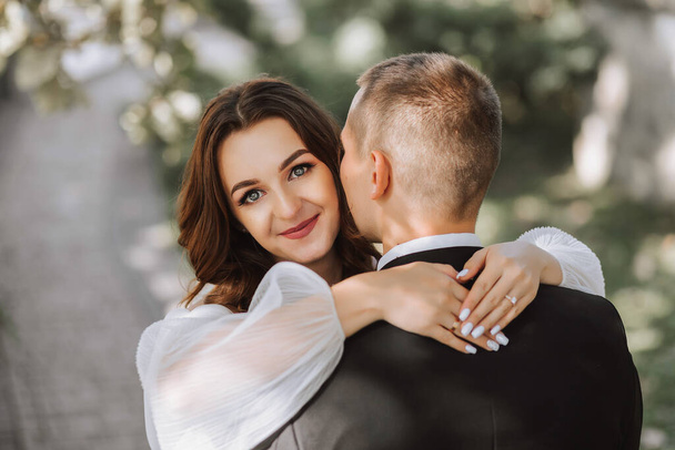 a girl hugs her boyfriend by the neck, the photo is taken from the back, beautiful female hands on the shoulders of a man. The girl looks at the camera, the man kisses his wife on the cheek.Free space - Foto, afbeelding