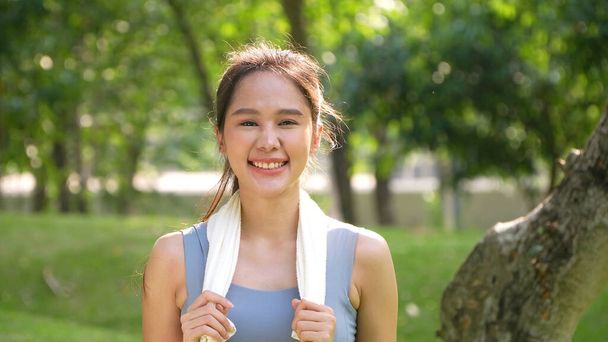 若いアジアの女性が魅力的な笑顔を描き、ワークアウト後に休んで白いタオルを使用します。アウトドアで働くスポーティな若い女性を笑顔とカメラを見て。健康的なライフスタイル｜健康 - 写真・画像