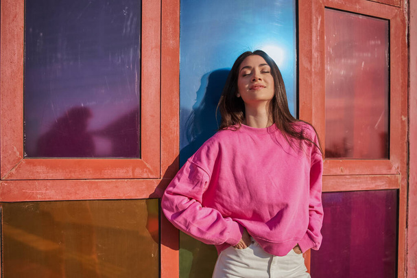 Trendy millennial woman in pink sweatshirt looking at the camera with light smile and posing at the sunny place - Foto, imagen
