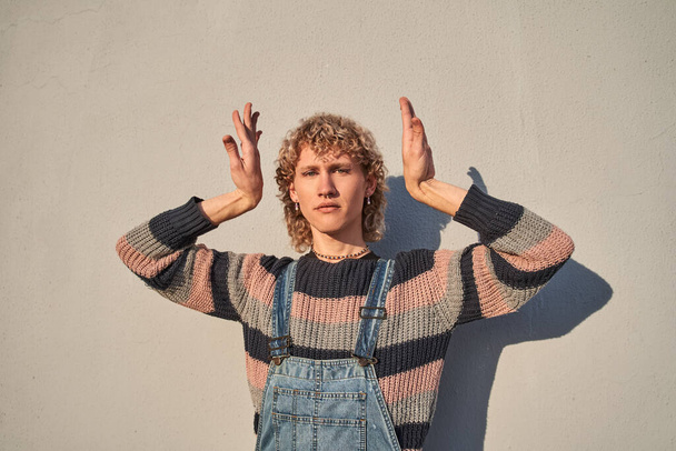 Trendy curly man standing with raised hands and looking at the camera with serious expression. Teen lifestyle concept - Foto, Bild
