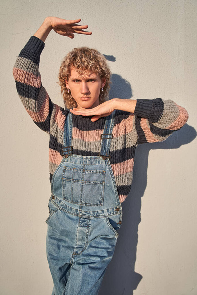 Vertical portrait of young man in trendy jeans overalls looking at camera with serious face and raised hands. People lifestyle concept - Foto, Bild
