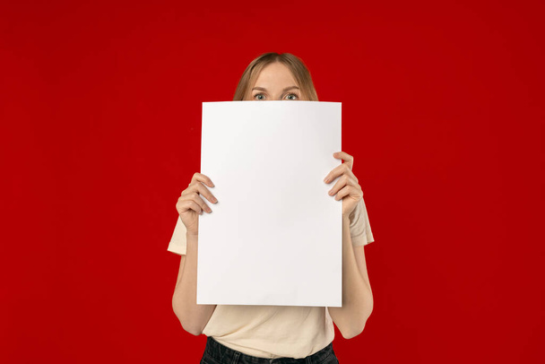 笑顔の白人女性は、赤いスタジオの背景に立って何かを宣伝空の垂直の白いポスターを保持.モックアップ - 写真・画像