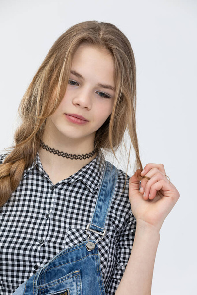 Bust of a girl posing for a photo on a white background. The girl is combed in a braid. She is wearing a plaid shirt. The shirt is buttoned up with black buttons. The girl is holding a strand of her - Fotografie, Obrázek