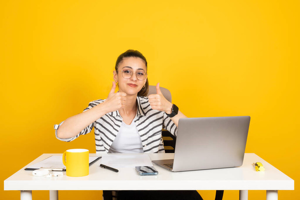 ジェスチャーのように親指を上げ、オフィスワーカービジネス女性の肖像画はジェスチャーのように親指を示すオフィスデスクで仕事に座る。黄色の背景が孤立。ラップトップを使用して、キャリアコンセプトを達成. - 写真・画像