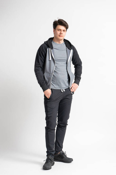 Young dark-haired man in a black tracksuit, gray t-shirt and running shoes posing full length in the studio on a white background - Photo, Image