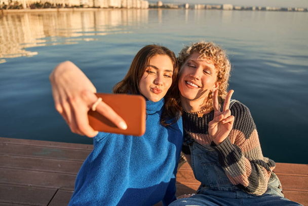 Positive teen couple gesturing hands and taking mobile phone selfie at the blue ocean background. Technologies and people concept - Φωτογραφία, εικόνα