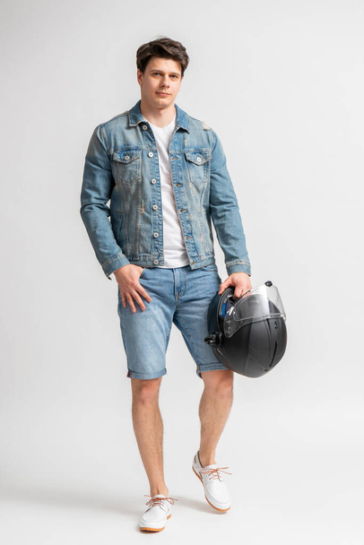 Young dark haired man in casual denim jacket and shorts, holding black protective motorcycle helmet, posing full length in studio on white background - Foto, imagen
