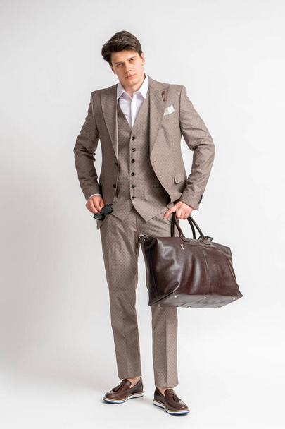 Молодой темноволосый мужчина в деловом костюме из трех частей с солнцезащитными очками и большой кожаной сумкой для путешествий и деловых поездок, позирующей в студии на белом фоне - Фото, изображение