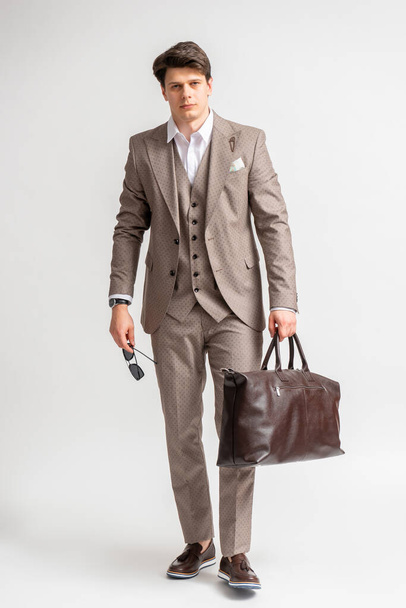 ビジネスの若いダークヘアの男サングラスと大きな革の旅行バッグ付きの3ピーススーツ旅行やビジネス旅行のための白い背景にスタジオで長さをポーズ - 写真・画像