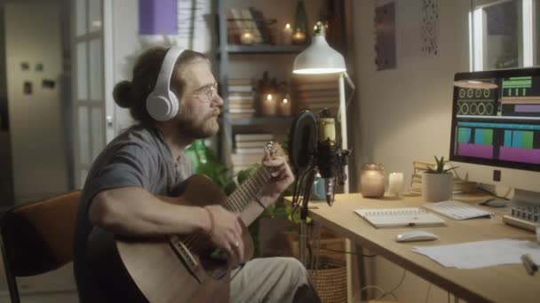 Grabación de arco medio del músico masculino en auriculares tocando la guitarra y cantando en el micrófono mientras graba la canción en el estudio en casa - Metraje, vídeo