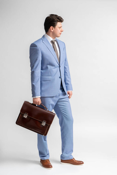 Młody, ciemnowłosy mężczyzna w niebieskim garniturze biznesowym, biała koszula i krawat ze skórzaną walizką w ręku pozuje w pełni w studiu na białym tle. - Zdjęcie, obraz