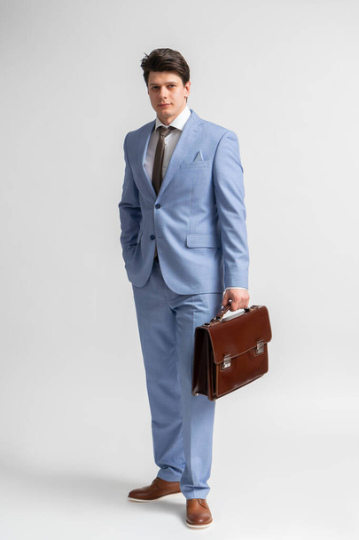 A young dark-haired man in a blue business suit, white shirt and tie with a leather briefcase in his hands poses in full growth in the studio on a white background - Fotografie, Obrázek