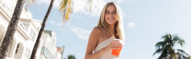 Młoda, piękna blondynka w Miami elegancko trzyma kubek orzeźwiającego drinka w białej sukience. - Zdjęcie, obraz