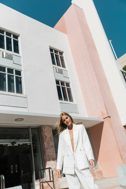 白いスーツの見事な若いブロンドの女性は,マイアミの近代的な建物の前に自信を持って立っています. - 写真・画像