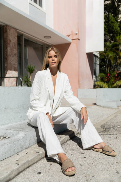 Una giovane donna serena con i capelli biondi, elegantemente vestita con un abito bianco, si siede aggraziatamente a terra a Miami. - Foto, immagini