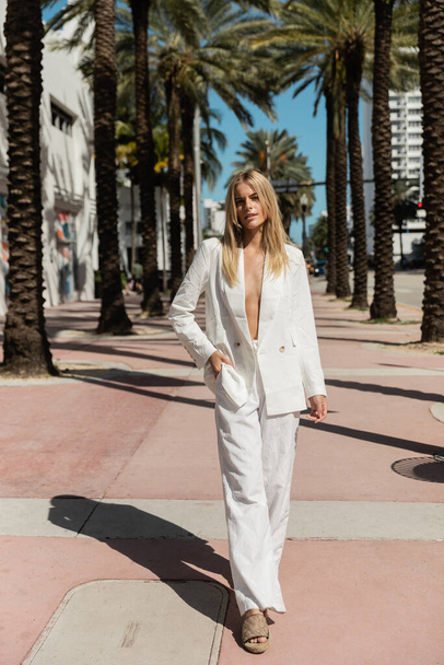 Una donna bionda trasuda fiducia mentre cammina lungo una strada di Miami in uno splendido abito bianco, una visione di eleganza. - Foto, immagini