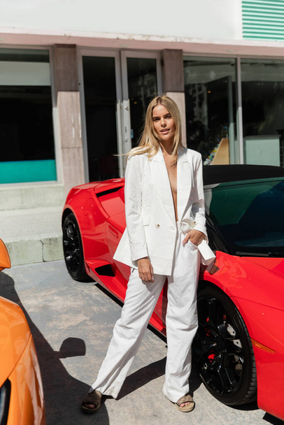 Una joven y hermosa mujer rubia de pie con confianza junto a un elegante coche deportivo rojo en Miami. - Foto, imagen