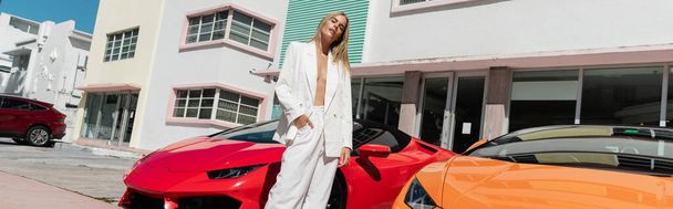Eine atemberaubende junge Frau mit blonden Haaren steht elegant neben einem leuchtend roten Sportwagen in Miami. - Foto, Bild