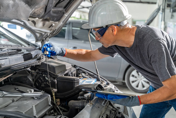 An Asian male car mechanic holding a computer tablet while checking car oil and working on the engine at his car repair garage - Foto, Bild