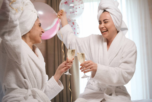 Lüks otel odasında bornoz giyen mutlu kadınların portresi ve köpüklü şarapla kadeh tokuşturma. - Fotoğraf, Görsel