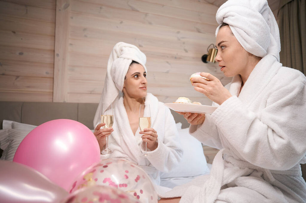 Cheerful females wearing bathrobe lying in big bed in hotel room while holding cakes and glasses with sparkling wine - Photo, Image
