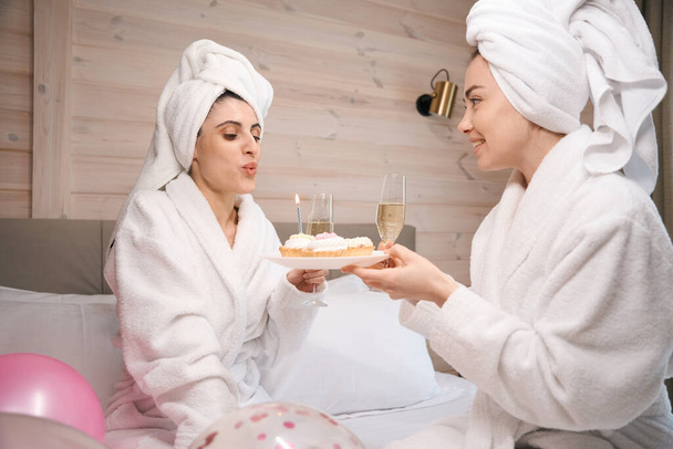 Glückliche Frauen im weißen Bademantel halten Gläser mit Sekt in der Hand und pusten Kerzen auf Kuchen aus. - Foto, Bild