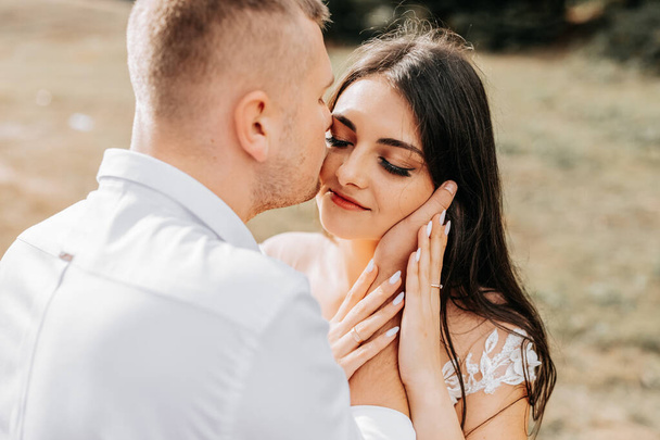 A stylish couple of European newlyweds. A smiling bride in a white dress hugs the groom from behind. The groom is dressed in a white shirt, without a tie. Outdoor wedding in the mountains - Φωτογραφία, εικόνα