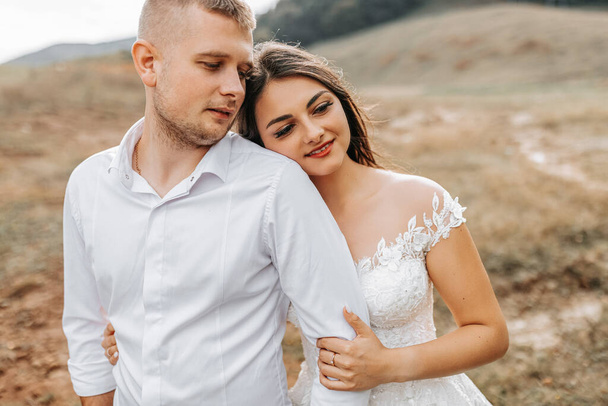 A stylish couple of European newlyweds. A smiling bride in a white dress hugs the groom from behind. The groom is dressed in a white shirt, without a tie. Outdoor wedding in the mountains - Fotoğraf, Görsel