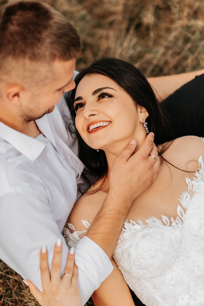 Groom and smiling bride lie on dry grass and hug, woman in white wedding dress. Beautiful autumn wedding photo. The groom aims at her forehead - Фото, зображення