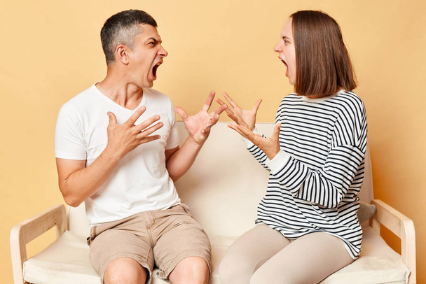 Irritatedannoyed woman and man dressedcasuallysitting on sofa isolated over beige background arguing at home screaming with hate and anger, relationship problems. - Fotografie, Obrázek