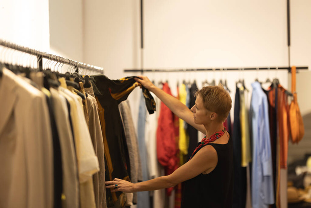 Ενηλίκων κομψό γυναίκα σύμβουλος μόδας ψάχνει πάνω από τα ρούχα στη μπουτίκ. Μεσαίο πλάνο - Φωτογραφία, εικόνα