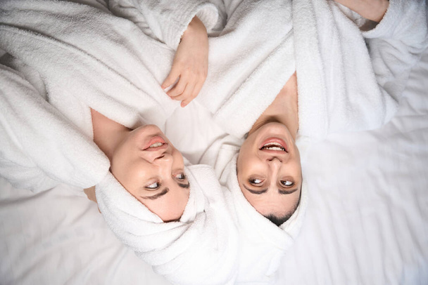 Beyaz bornozlu iki kadının portresi. Vücut tedavisinden memnun olarak yatakta yatıyorlar. - Fotoğraf, Görsel