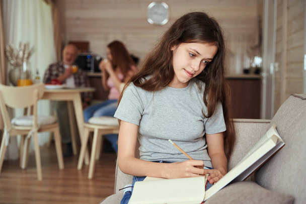 Calma concentrato homeschooler seduto sul divano a fare i compiti mentre i genitori bevono succo d'arancia al tavolo della cucina - Foto, immagini