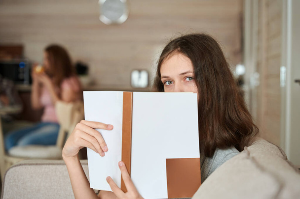 Jovencita sentada en un sofá en casa mientras esconde su cara detrás de un libro de texto abierto - Foto, Imagen