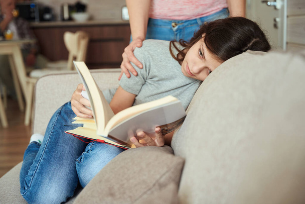 Jovencita concentrada tumbada en el sofá mientras lee el libro en presencia de la madre - Foto, imagen