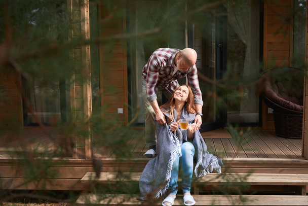 Caring husband draping plaid around shoulders of his smiling pleased wife seated on porch - Photo, Image