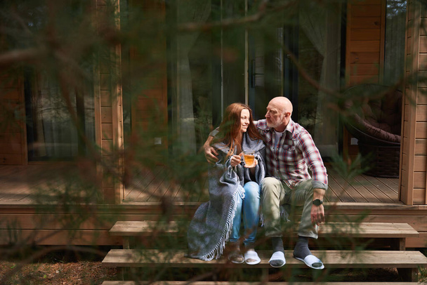 Amado marido abraçando esposa sorridente com xícara de chá em mãos sentadas ao lado dele em degraus varanda de madeira - Foto, Imagem