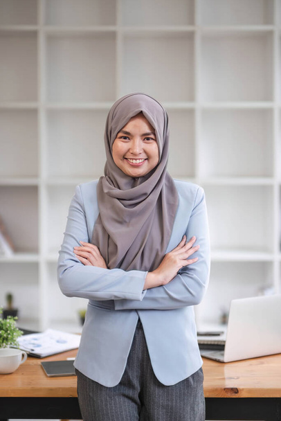 A portrait of a beautiful and professional Asian Muslim businesswoman stands in the office with her arms crossed... - Foto, imagen