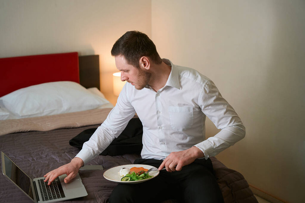 Σοβαρός νεαρός που κάθεται στο κρεβάτι στο δωμάτιο του ξενοδοχείου τρυπώντας το φαγητό με πιρούνι και πληκτρολογώντας στο πληκτρολόγιο laptop - Φωτογραφία, εικόνα