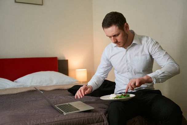 Αρσενικό τσίμπημα τροφίμων με πιρούνι, ενώ κάθεται στο κρεβάτι μπροστά από φορητό υπολογιστή σε aparthotel - Φωτογραφία, εικόνα