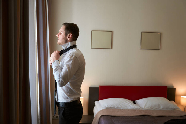 Zijaanzicht van elegante jongeman koppelverkoop stropdas rond shirt kraag terwijl kijken uit door gordijnen in luxe suite - Foto, afbeelding