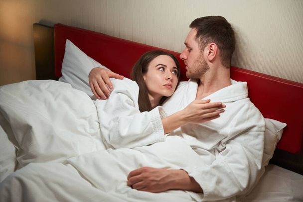 Homme étreignant son compagnon féminin sérieux couché sur son épaule au lit dans la chambre d'hôtel pendant la conversation - Photo, image