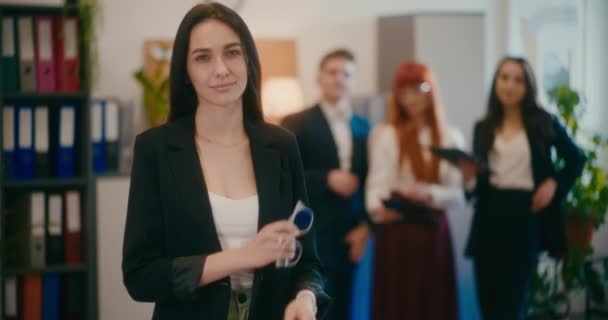 Портрет усміхненої молодої бізнес-леді, що стоїть зі зброєю, схрещеною, поки співробітники на задньому плані в офісі. - Кадри, відео