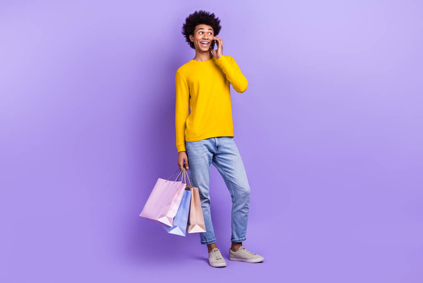 Full length photo of cheerful handsome man speak telephone hold store mall bags isolated on violet color background. - 写真・画像