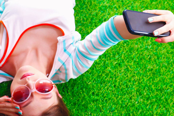 Beautiful young woman making selfie by her phone while lying in green grass. - Фото, изображение