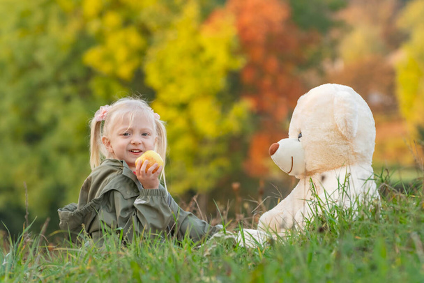 Blonde girl eats apple and plays with large soft toy sitting on grass. Child on walk on autumn day in nature. - Photo, Image