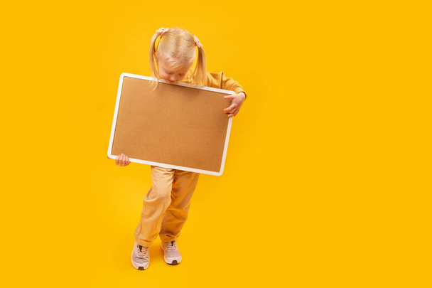 Klein blond meisje met blanco kurkbord op gele achtergrond. Ruimte voor je tekst. Kopieerruimte - Foto, afbeelding