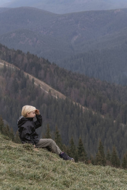 Child on vacation in mountain. Blond schoolboy sits at hillside and looks at the distance. Vertical frame. - Foto, afbeelding