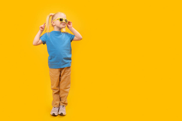 Feliz niña rubia divertida usar gafas de sol y camiseta azul sobre fondo amarillo. Copiar espacio, burlarse. - Foto, Imagen