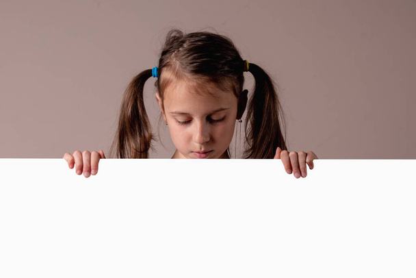 Pretty girl looking down behind empty whiteboard. Child holding paper poster advertising Your text. - Foto, Imagen